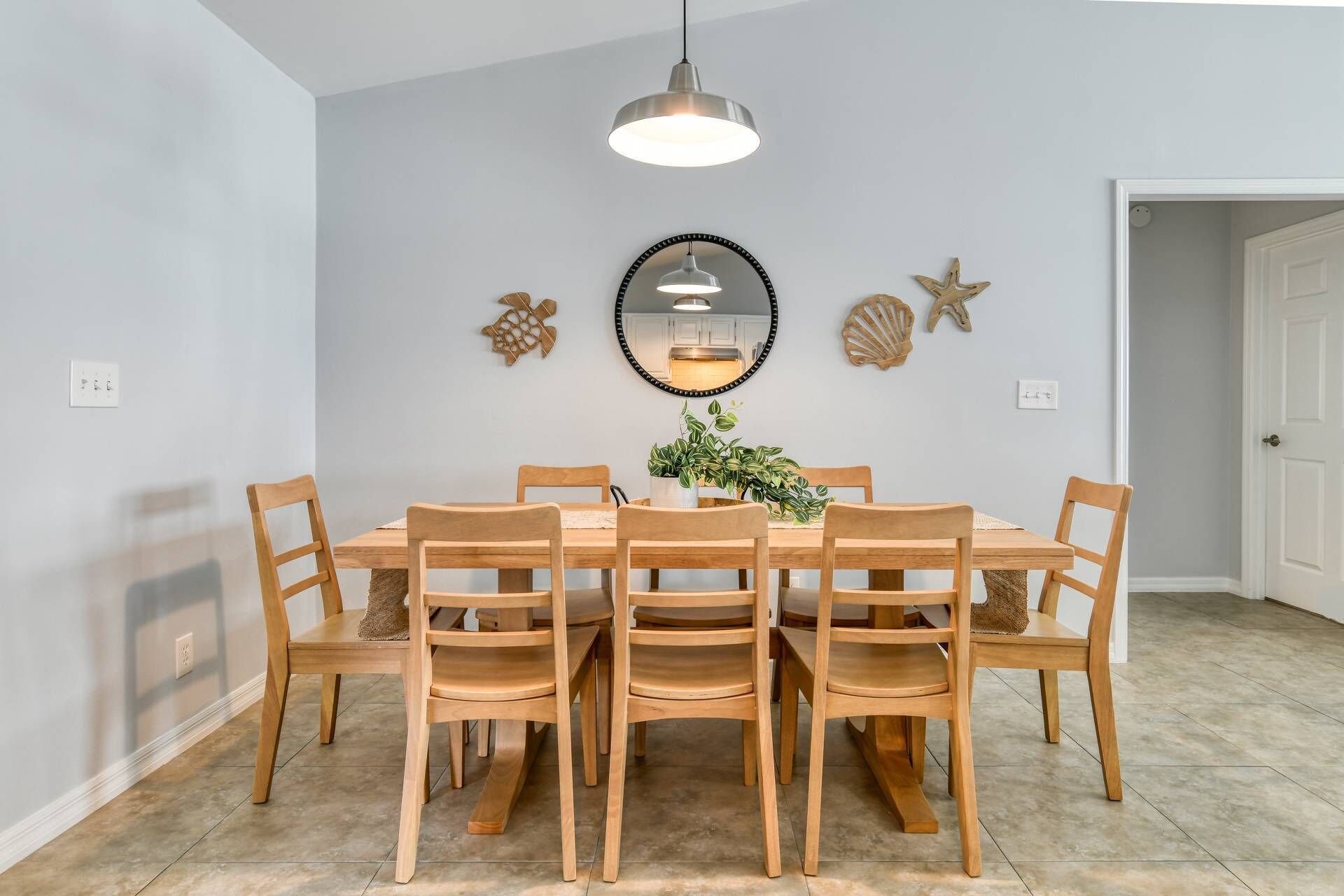 A dining room with a round table, six wooden chairs, a hanging pendant light, a mirror, and decorative shells on the wall.
