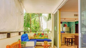 A balcony with a sofa, a small table, and chairs. There are plants and a view of a building with a green wall and a kitchen area.