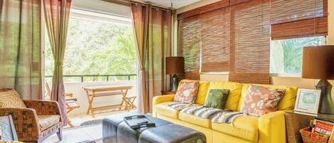 A living room with a yellow sofa, a black ottoman, and a patterned armchair. There is a ceiling fan, a window with curtains, and a view of greenery outside.