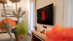 Un salon moderne avec un téléviseur à écran plat fixé au mur, un banc en bois, une plante en pot et une petite table sur laquelle se trouvent un panier et un téléphone.