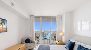 A hotel room with a bed, a chair, a desk, a lamp, and a view of a cityscape through large windows.