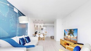 A modern living room with a white sofa, blue cushions, a dining area with a white table and chairs, a kitchen with white cabinets, and a TV on a wooden stand. - Upper Apartment: Pool + Gym + Amazing view (Hallandale Beach)