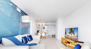 Una sala de estar moderna con un sofá blanco, cojines azules, una zona de comedor con una mesa y sillas blancas, una cocina con armarios blancos y un televisor sobre un mueble de madera.