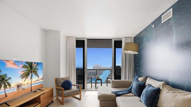 A modern living room with a large window, a sofa, a chair, and a TV. - Upper Apartment: Pool + Gym + Amazing view (Hallandale Beach)
