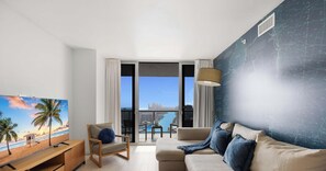 A modern living room with a large window, a sofa, a chair, and a TV. - Upper Apartment: Pool + Gym + Amazing view (Hallandale Beach)