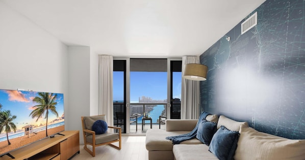 A modern living room with a TV, a sofa, a chair, and a balcony with a view of the city and water.