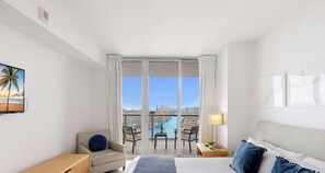 A hotel room with a bed, a chair, a desk, a lamp, and a view of a cityscape through large windows.