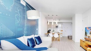 Een moderne woonkamer met een witte bank, blauwe kussens, een eethoek met een witte tafel en stoelen, een keuken met witte kastjes, en een tv op een houten dressoir.
