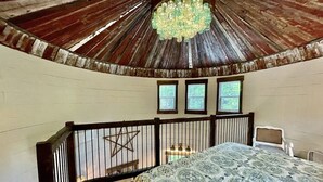 A bedroom with a wooden ceiling, a bed with a patterned bedspread, a small table, and a chair. There are three windows and a star decoration on the wall.