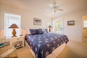 Une chambre à coucher comprenant un lit, deux tables de chevet, une commode, une chaise, un ventilateur de plafond et une porte donnant sur un espace extérieur.
