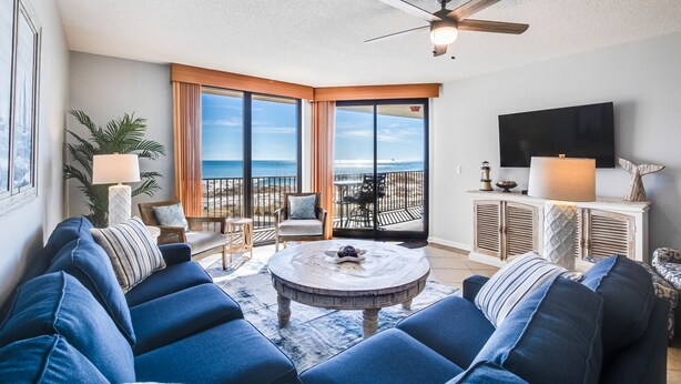 A spacious living room with a large sofa, a coffee table, and a ceiling fan. There is a balcony with a view of the ocean. - Perfect location at Phoenix X next door to FloraBama (Orange Beach)