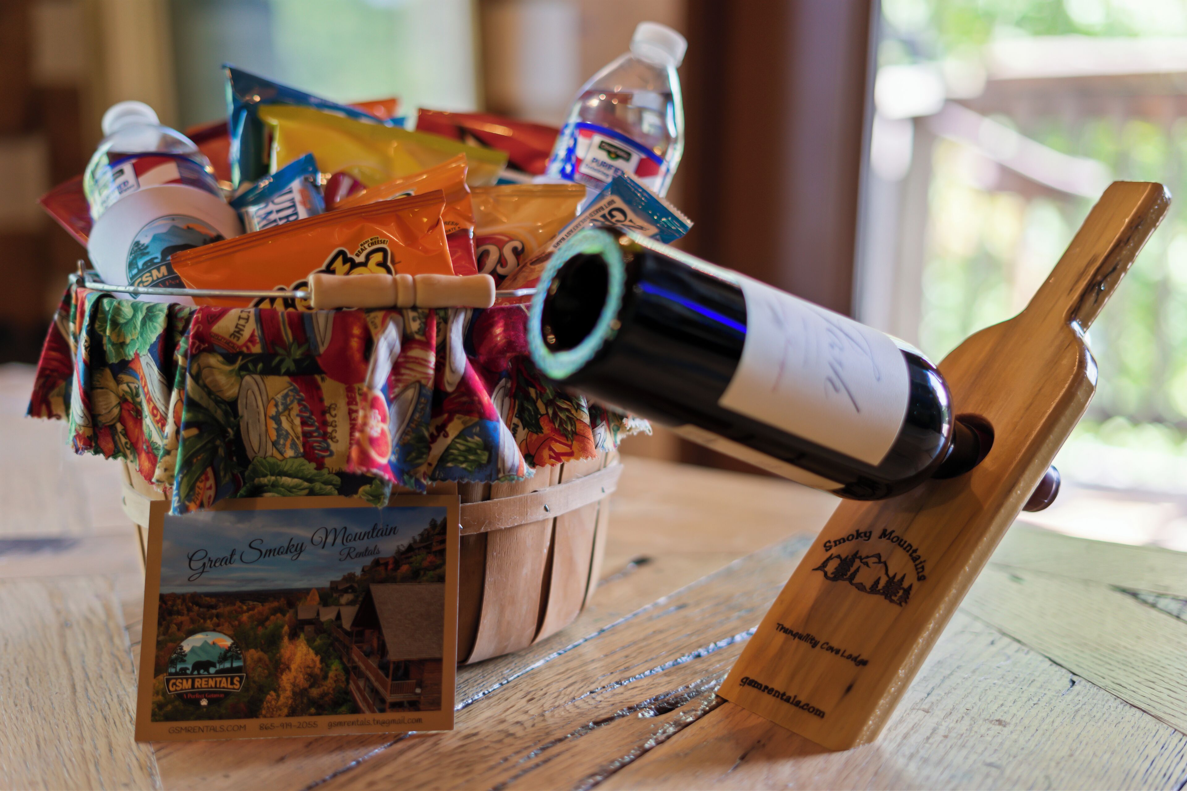 A wooden table with a basket containing snacks, a bottle of water, a bottle of wine, and a wooden wine opener.
