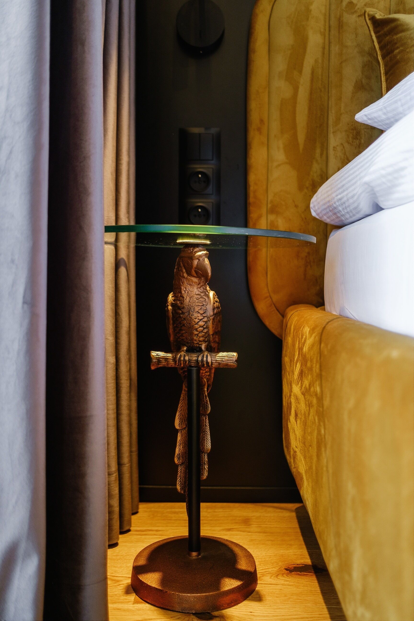 Una stanza con pavimento in legno, un letto con testiera bianca, un tavolino con piano in vetro e un oggetto decorativo a forma di uccello.