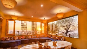 Una habitación acogedora con una mesa de madera, asientos acolchados y grandes ventanales con vistas a árboles y montañas.