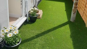 A narrow pathway with artificial green turf, potted plants, and a brick wall.