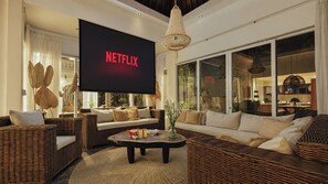 Un soggiorno con un'ampia TV a schermo piatto che mostra 'NETFLIX', mobili in vimini, un tavolino con oggetti e vista sulla cucina.
