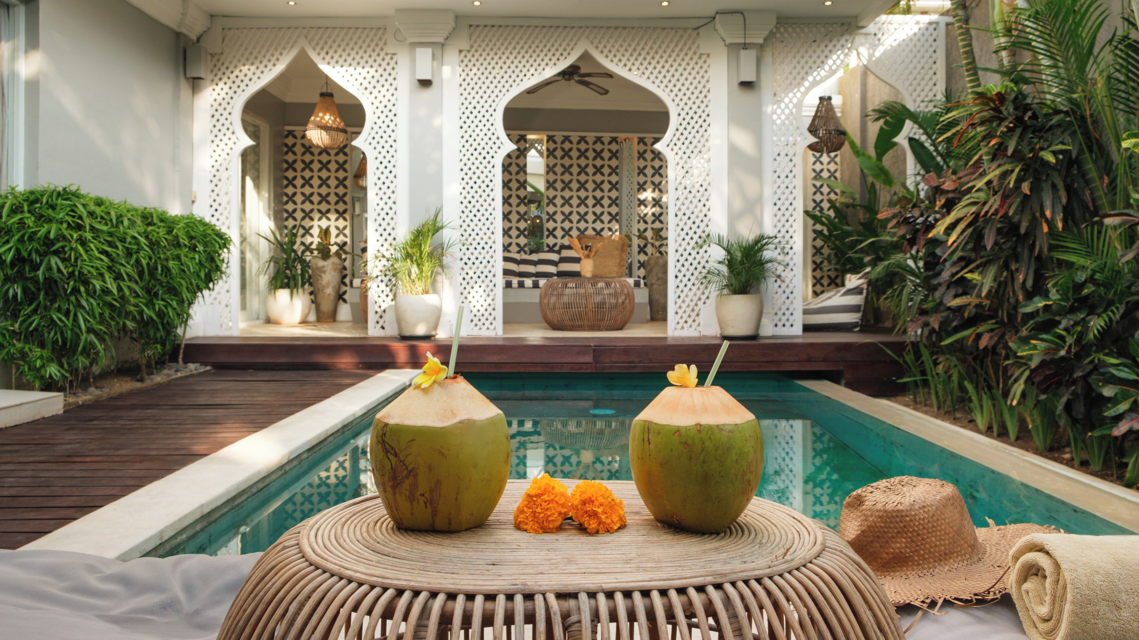 A pool area with two coconuts on a woven table, surrounded by lush greenery and a patterned wall.