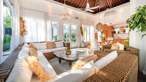 A spacious living area with a large sofa, a coffee table, and wicker furniture. There are large windows with curtains, a ceiling fan, and a view of the outdoors. - Villa Moana Canggu by Villa Finder (Canggu)