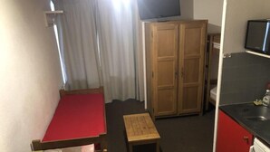 Una habitación pequeña con cama, una mesita, un armario de madera y una zona de kitchenette.