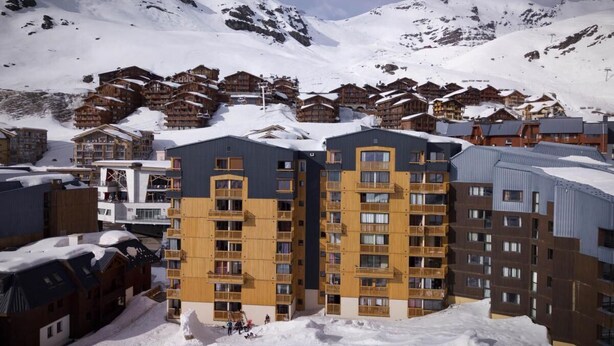Un complejo turístico en la montaña cubierto de nieve, con edificios de varios pisos que tienen balcones y un telesilla al fondo.