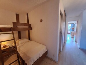 A single bed in a room with a wooden bunk bed structure to the left. A door leads to a hallway with a view of a balcony.