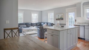 Een ruime woonkamer met een donker leren bank, een witte salontafel en een fauteuil met een patroon. Een keuken met witte kasten en een marmeren aanrecht is zichtbaar.