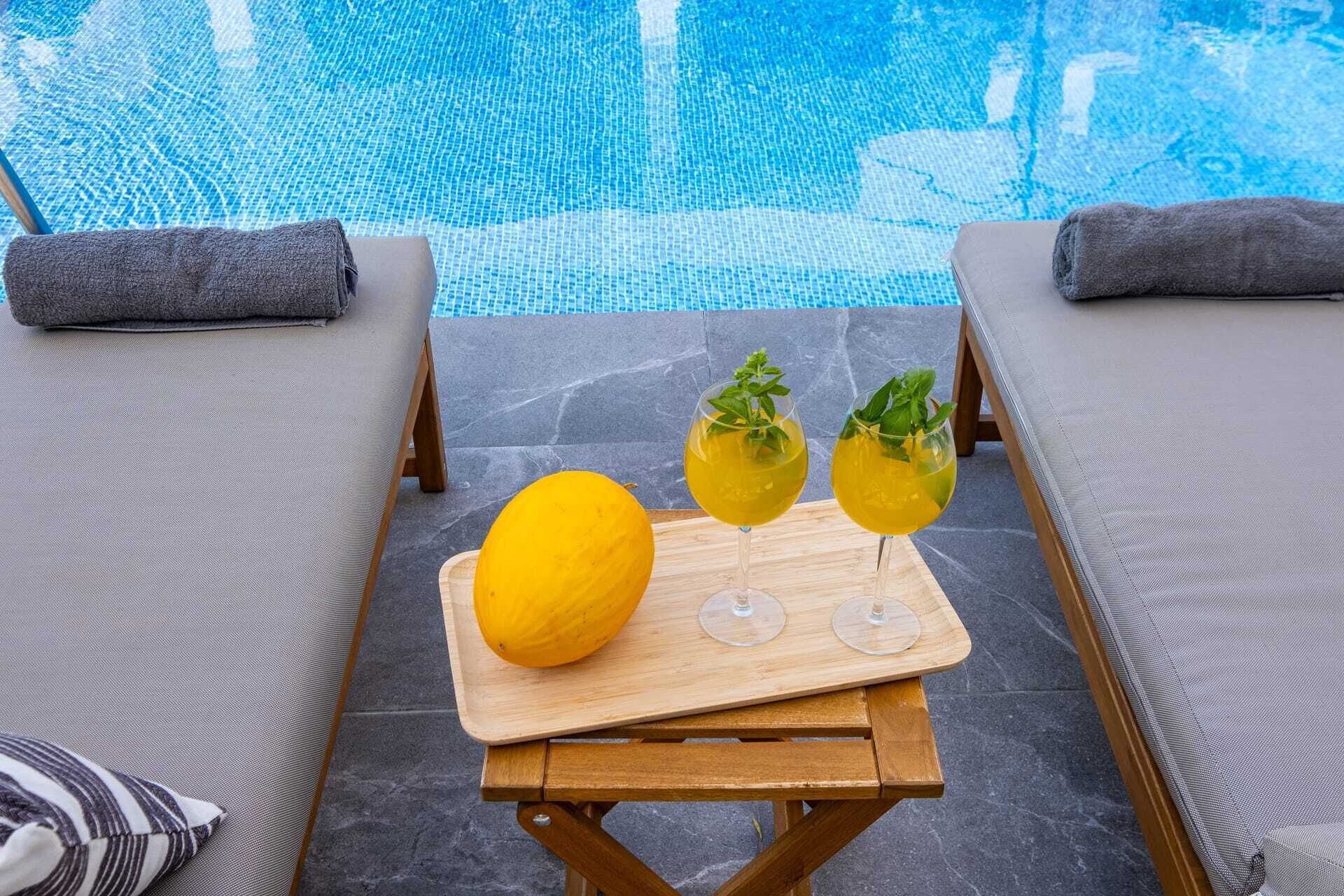Un espace aménagé au bord de la piscine, avec des chaises longues, une table en bois sur laquelle se trouvent un citron et deux verres remplis d’une boisson jaune, et une vue dégagée sur la piscine.