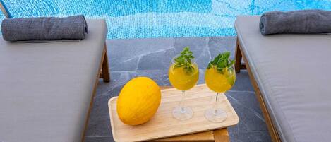 Un espace aménagé au bord de la piscine, avec des chaises longues, une table en bois sur laquelle se trouvent un citron et deux verres remplis d’une boisson jaune, et une vue dégagée sur la piscine.