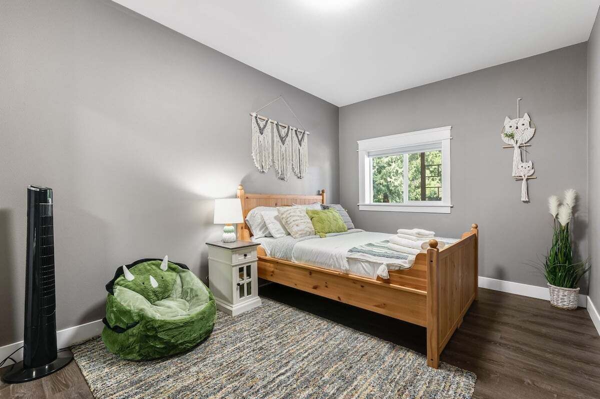 A bedroom with a wooden bed, a bedside table, a green bean bag chair, a standing fan, and a window with a view of greenery.