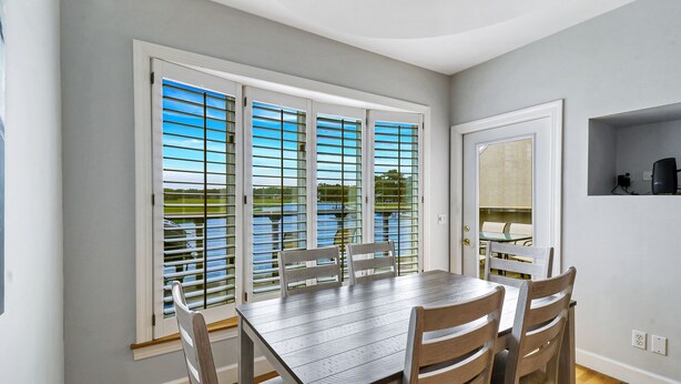 A dining area with a table and chairs, large windows with blinds, a door, and a small shelf.