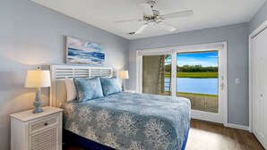 A bedroom with a bed, nightstands, a ceiling fan, a painting, and a view of a lake through a sliding door.