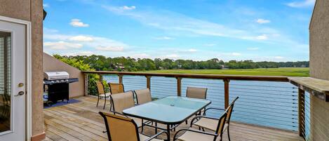 A deck with a table and chairs, overlooking a body of water and a park.