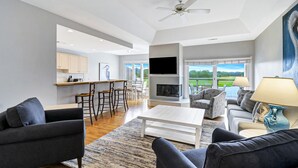 A spacious living area with a fireplace, comfortable seating, a kitchen with bar stools, and large windows offering a view of the outdoors.