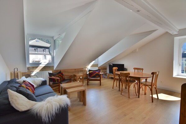 Un espace de vie de style loft avec un plafond haut, un plancher en bois, un canapé agrémenté d’un plaid en fausse fourrure, une table à manger avec des chaises et une vue sur une maison, visible à travers une lucarne.