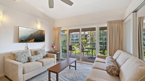 A hotel room with a sofa, a coffee table, a TV mounted on the wall, and a balcony with a view of buildings and greenery. - Kailani #314, Top Floor, Walk to Kapaa Town and Beach (Kapaa)