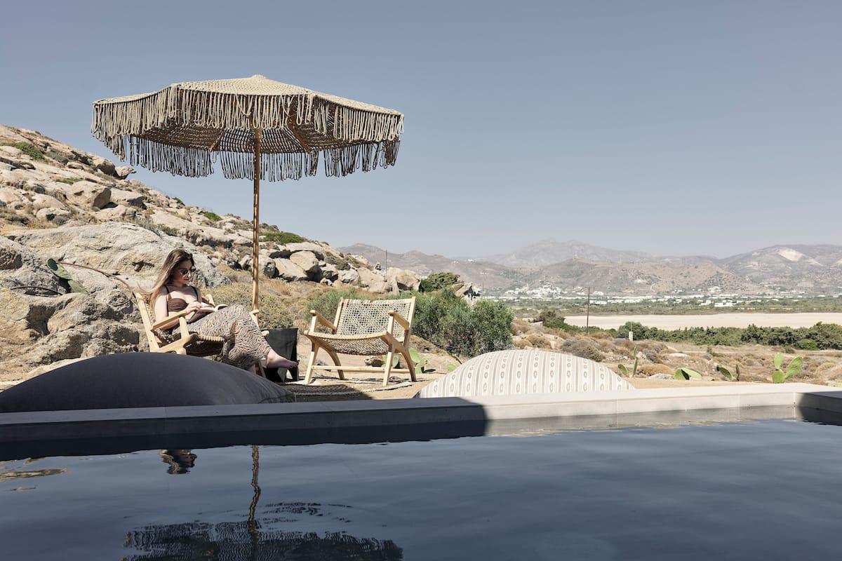 A pool area with two lounge chairs and a woven umbrella, surrounded by rocky terrain and distant mountains.