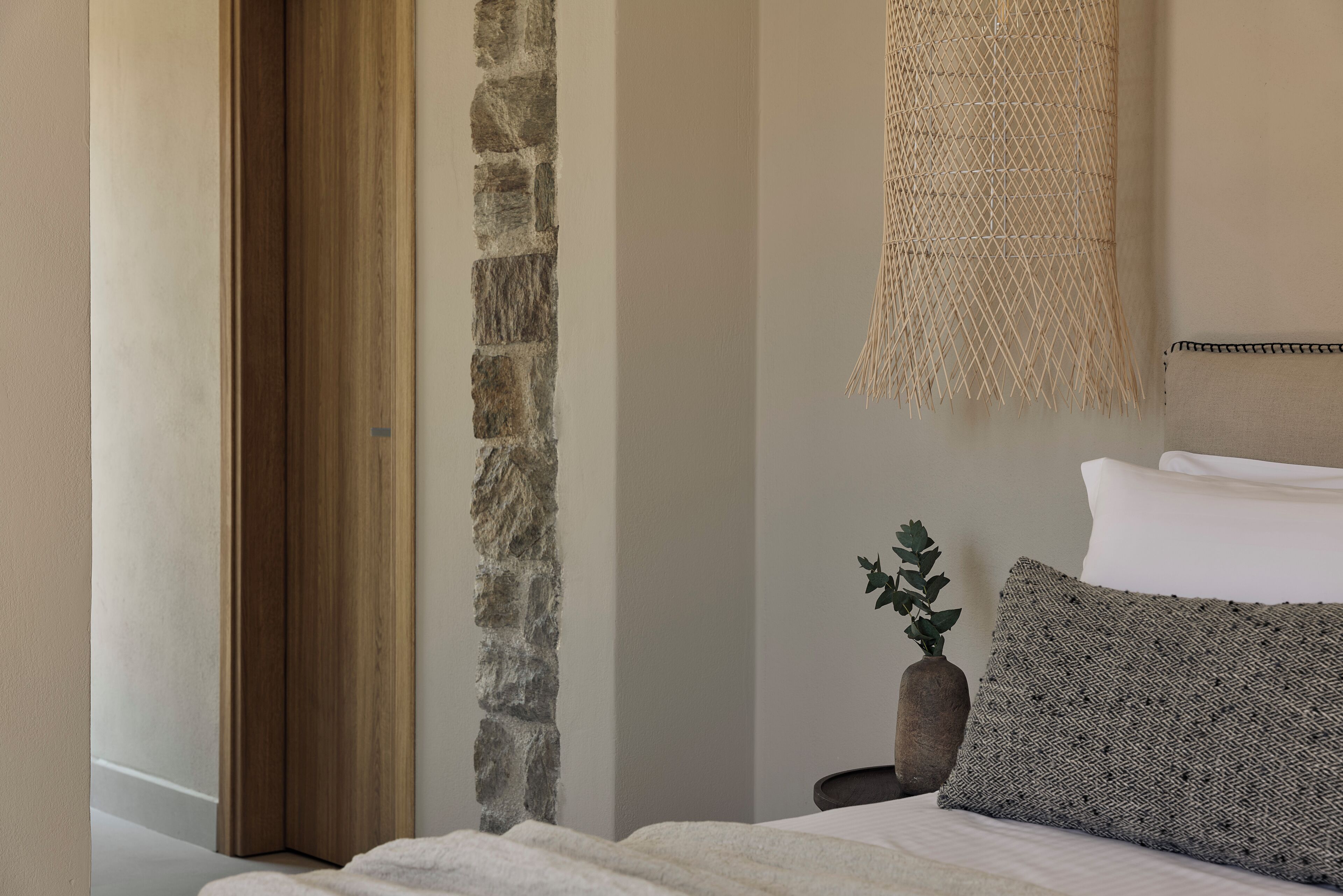 A bedroom with a stone wall, a woven headboard, a bed with white linens, and a small plant on a nightstand.