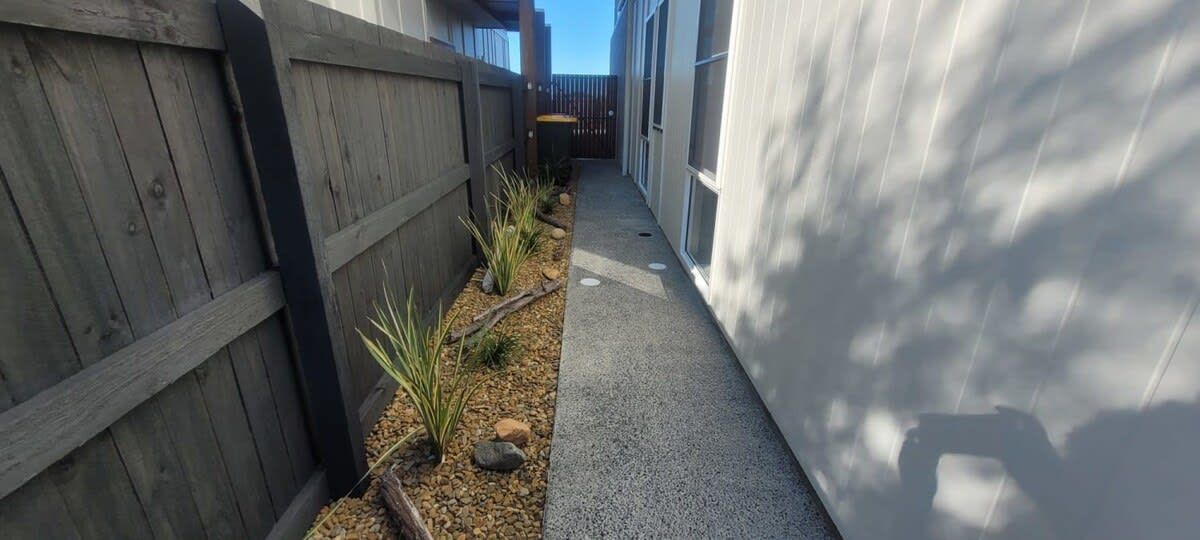 A narrow concrete pathway flanked by a wooden fence and a building with large windows.