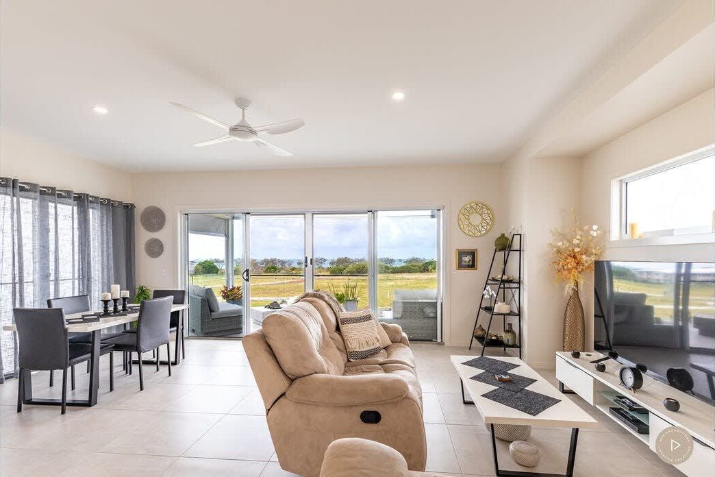 A modern living room with a sofa, dining table, chairs, a TV, and a balcony with a view of the countryside.