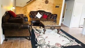 A cozy living room with a brown leather sofa, a matching armchair, and a wooden coffee table. A wooden staircase leads to an upper level. - Newly Remodeled home with Class A Trout Stream (Austin)