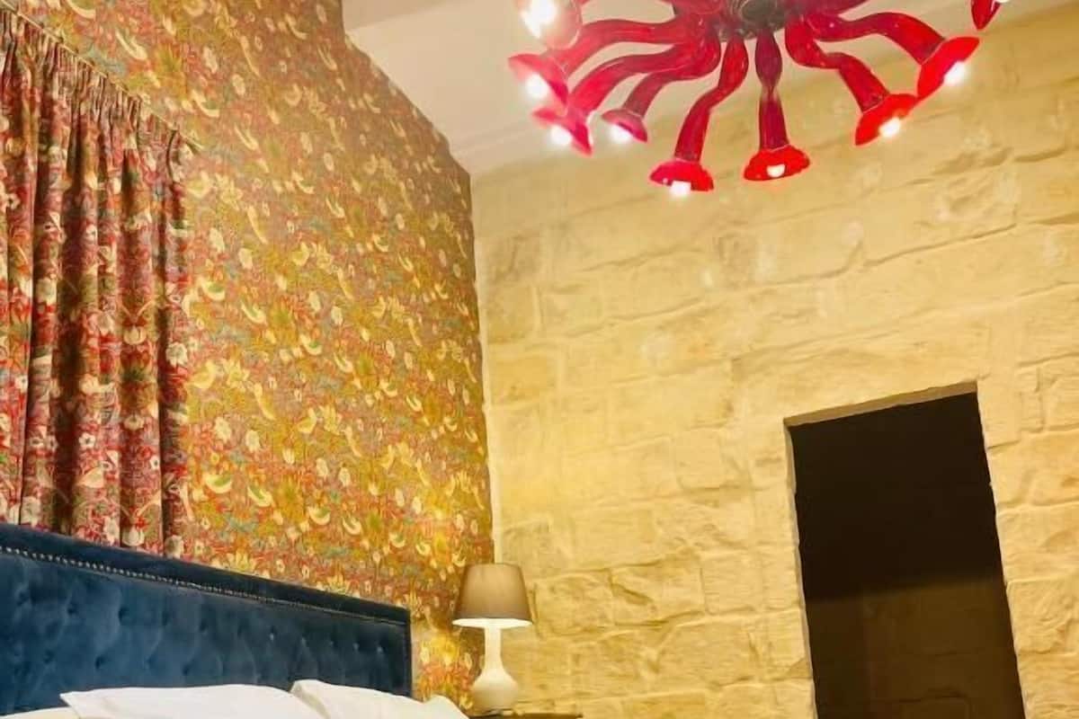 A bedroom with a bed, a red blanket, a wall with a floral pattern, a stone wall, and a unique ceiling light fixture.
