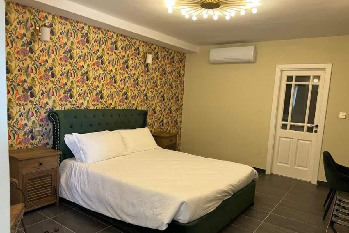 A bedroom with a green upholstered bed, a floral-patterned wallpaper, a white door, and a ceiling light fixture.