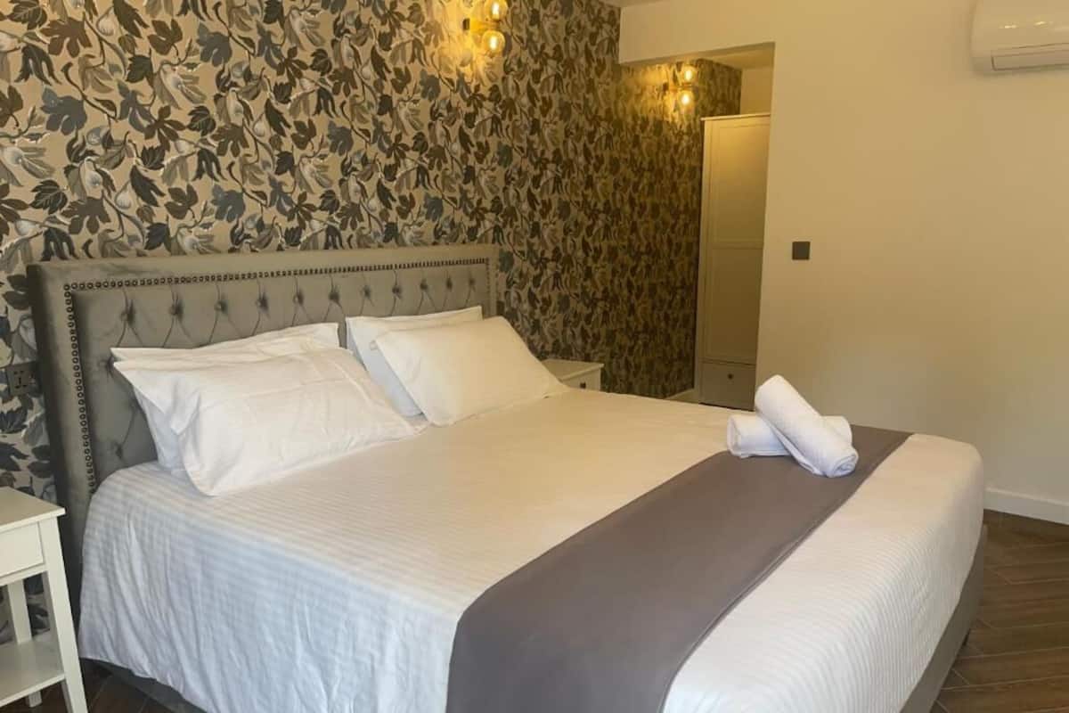 A neatly made bed with white linens and a gray accent, a white nightstand, and a floral-patterned wallpaper.