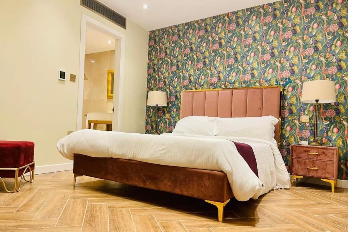 A bedroom with a large bed, a red bench, a nightstand, and a floral wallpaper.