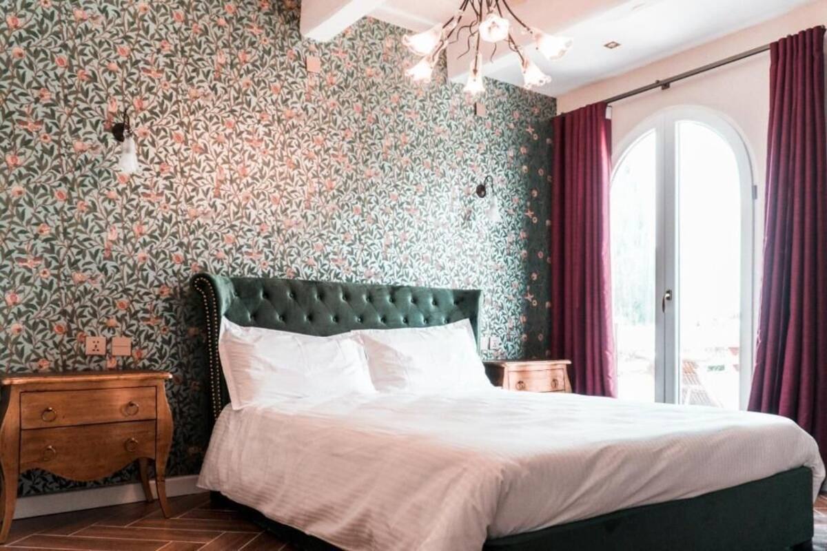 A bedroom with a floral wallpaper, a green upholstered headboard, a wooden nightstand, and a large window with curtains.