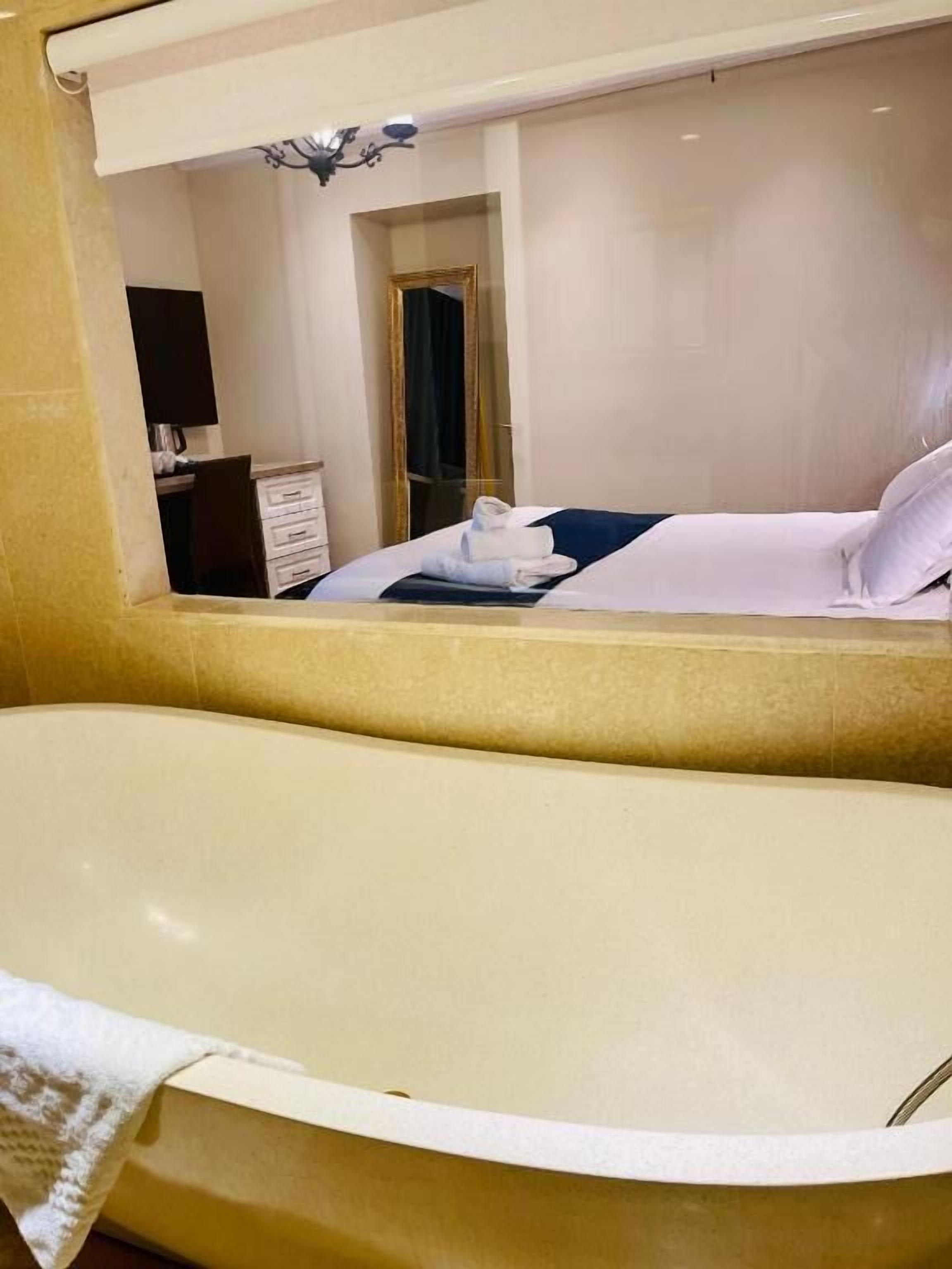 A hotel bathroom with a large bathtub, a mirror reflecting a bed and a door, and a chandelier.