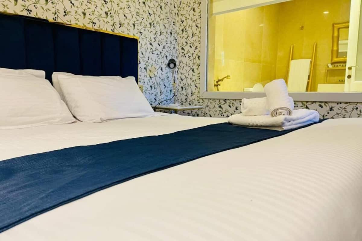 A neatly made bed with white linens, a dark headboard, and a window with a view of the bathroom.