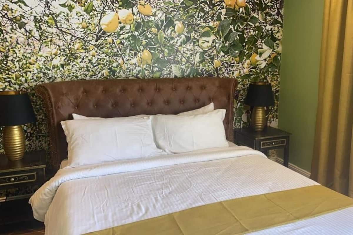 A neatly made bed with white linens and a gold accent, flanked by two bedside tables with lamps, in a room with a floral wallpaper backdrop.