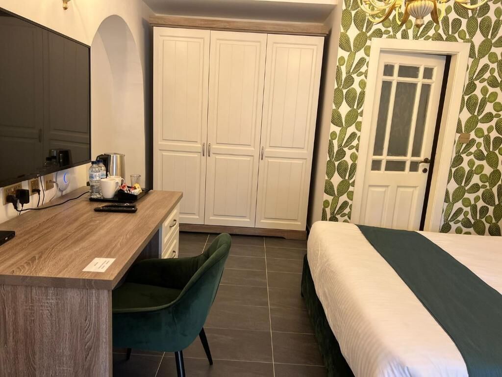 A hotel room with a wooden desk, a green chair, a bed with a green and white bedspread, a wardrobe, and a door with a cactus-patterned wallpaper.