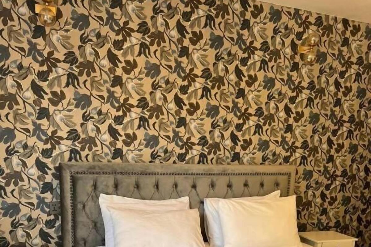 A neatly made bed with white linens and a grey headboard in a room with floral wallpaper.